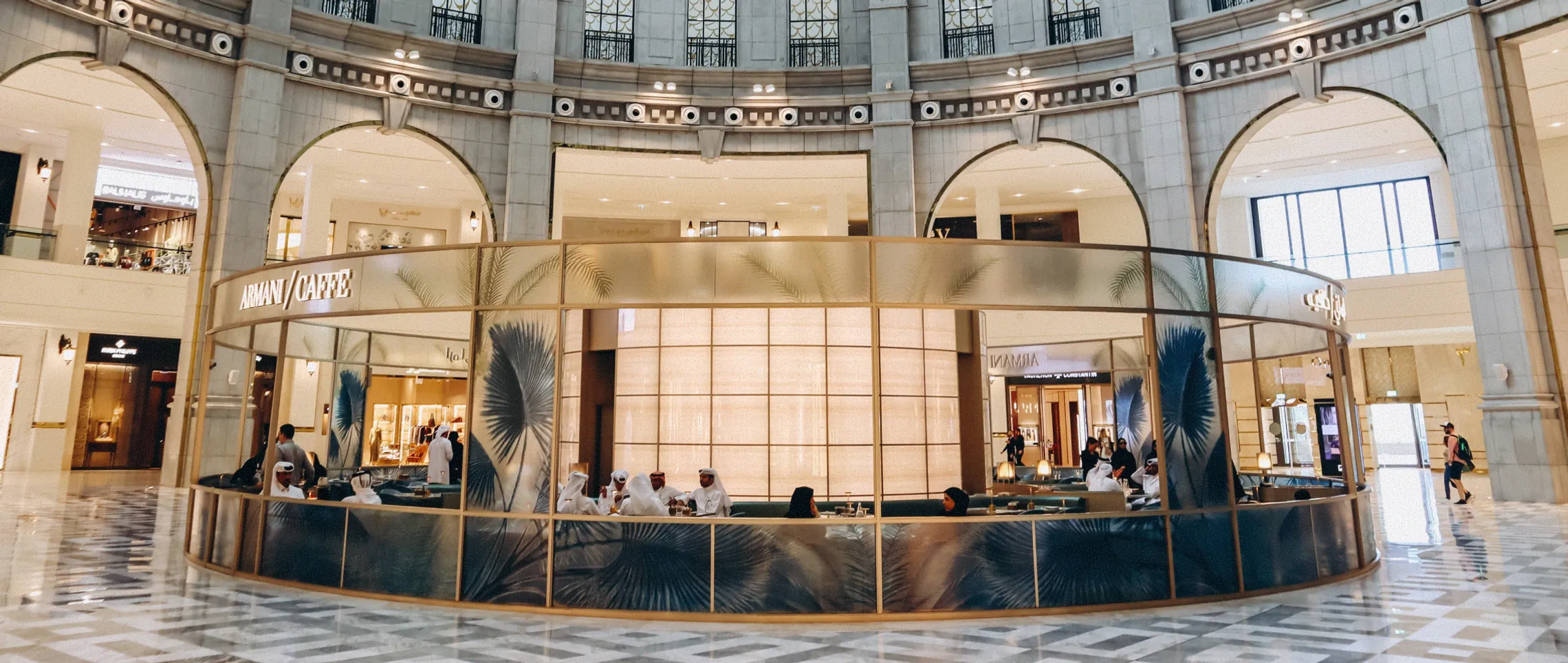 People seated inside a circular Armani Caffé in a spacious mall with marble floors and arches.